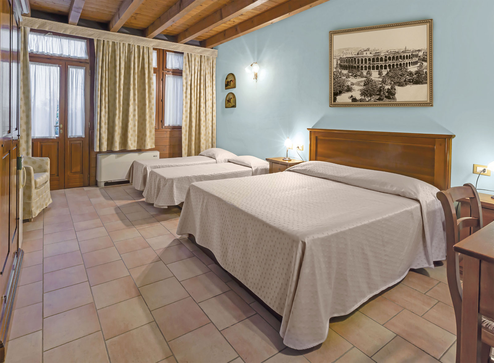 Vista panoramica di una delle camere dell'agriturismo casa rosa: travi a vista, colore azzurro delle pareti, letto matrimoniale e due letti singoli, ampia e spaziosa con armadio, potrona e scrivania. Luminosa dalle ampie finestre